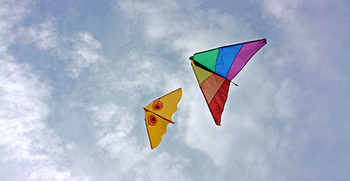 Two kites, one rainbow-striped and one yellow, flying against a cloudy sky